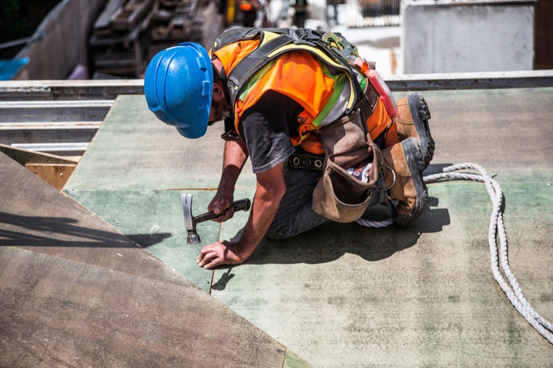 Roofer in safety gear hammering shingles on a rooftop